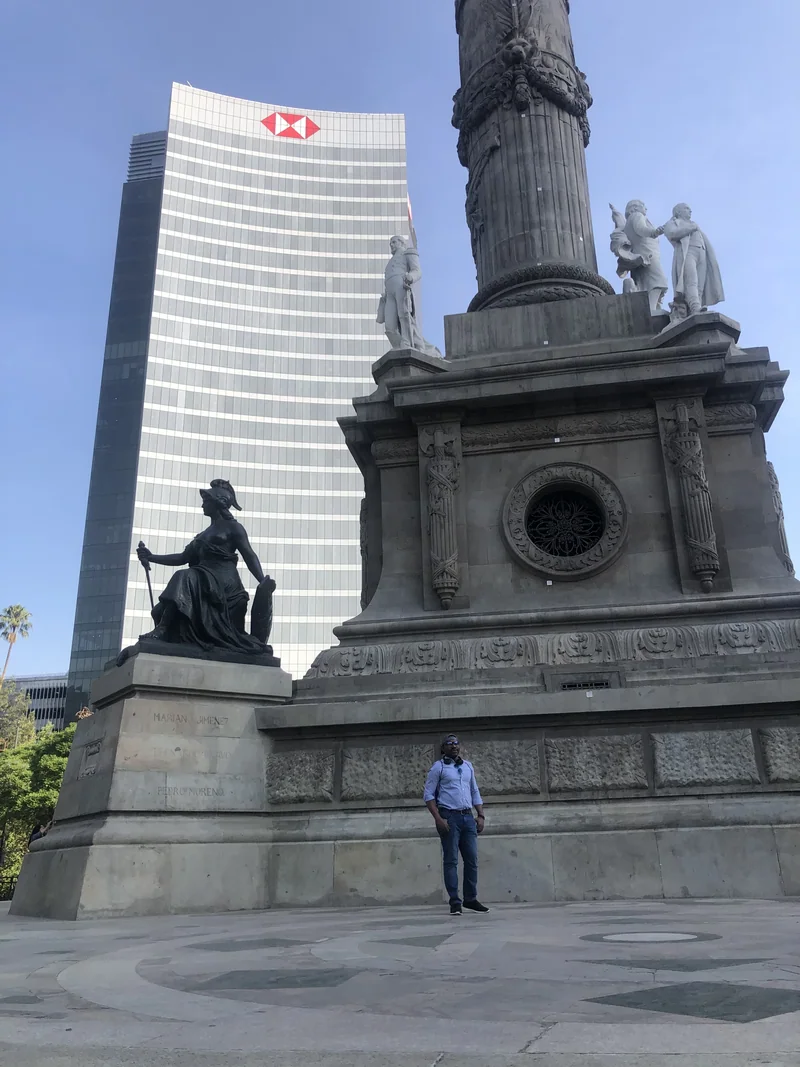A visitor stands beneath the carved pedestal and statues at the Ángel de la Independencia, with a glass tower in the background.