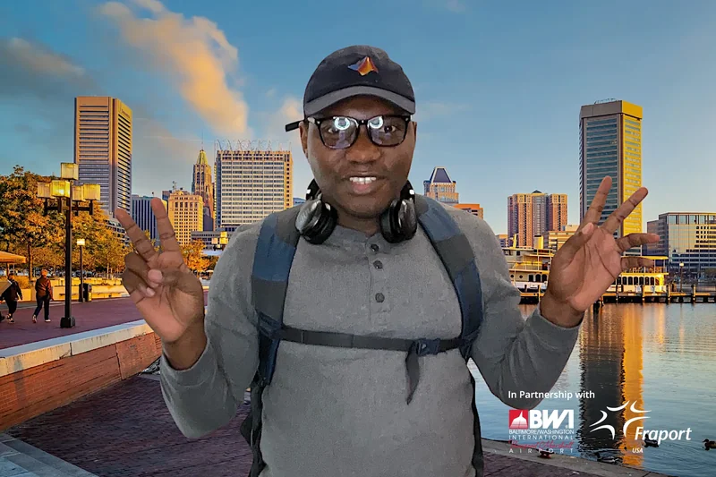 A traveler wearing a cap, headphones and backpack poses with both hands raised against a Baltimore waterfront photo-booth backdrop.