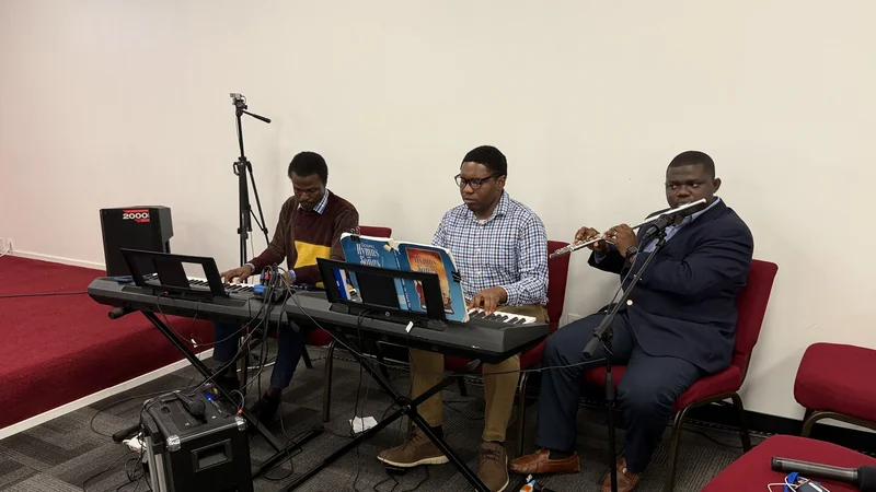 Three seated musicians perform together on two electronic keyboards and a flute, with hymn books open on a music stand.
