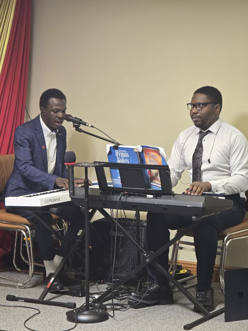 Two seated musicians play electronic keyboards; one sings into a microphone beside an open gospel hymn book.