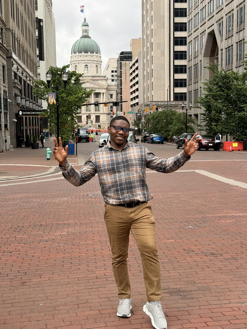 A visitor poses on a brick street, with the green dome of the Indiana Statehouse visible between downtown buildings.