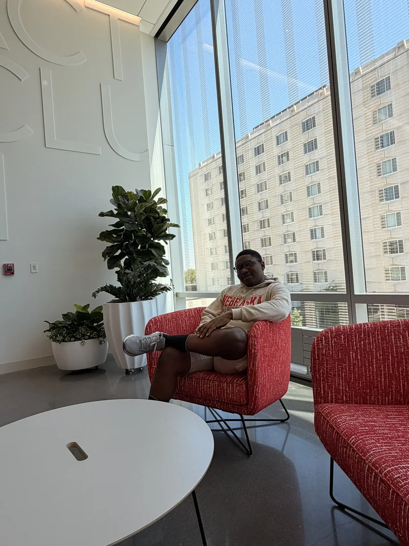 A student in a Nebraska sweatshirt sits in a red armchair beside tall windows and potted plants.