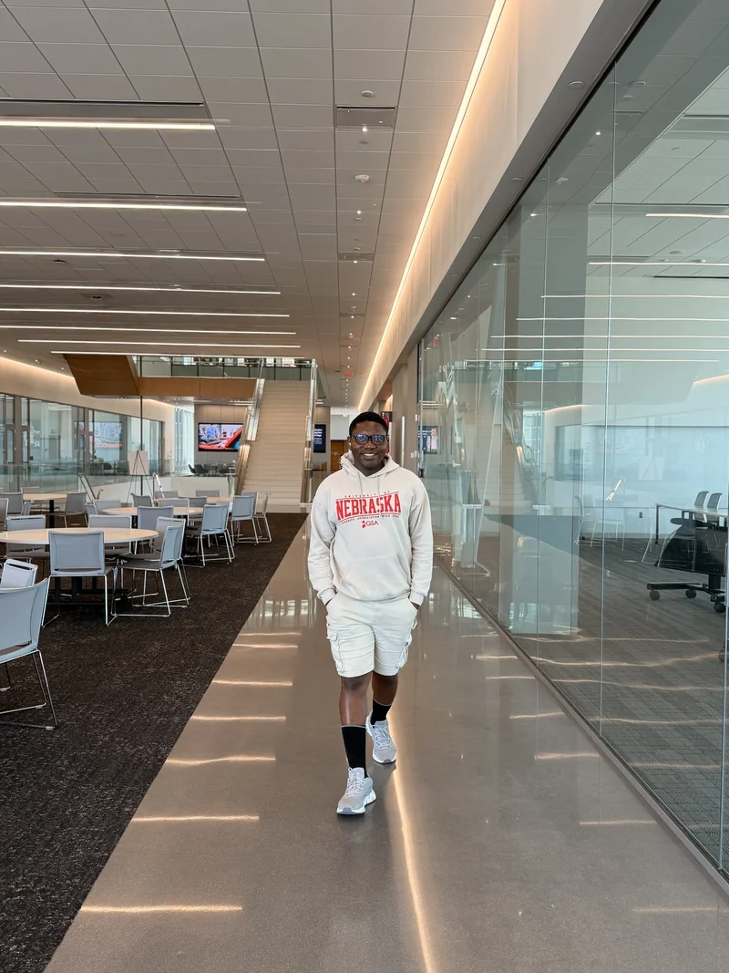 A student wearing a Nebraska sweatshirt stands in a bright glass-walled university commons with tables and a staircase behind.