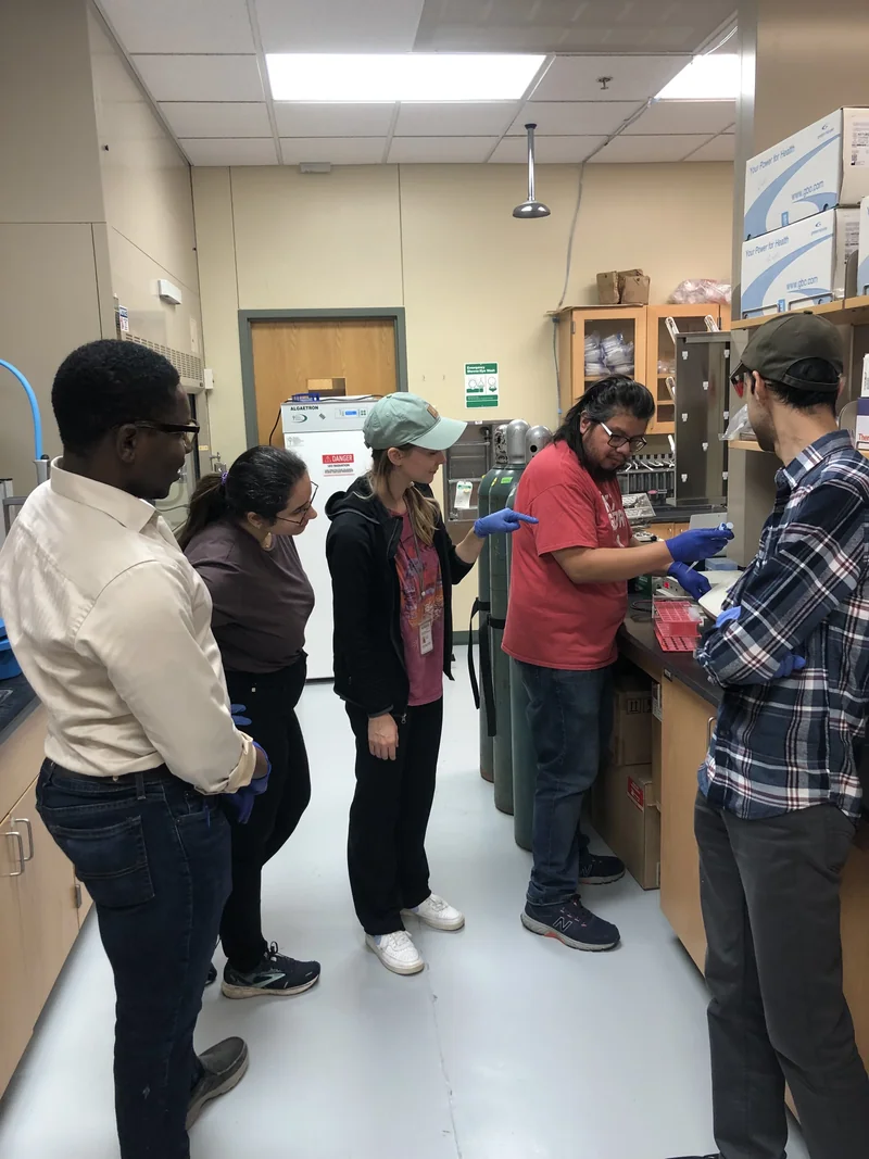 Five people stand around a laboratory bench, watching a colleague work with a micropipette.
