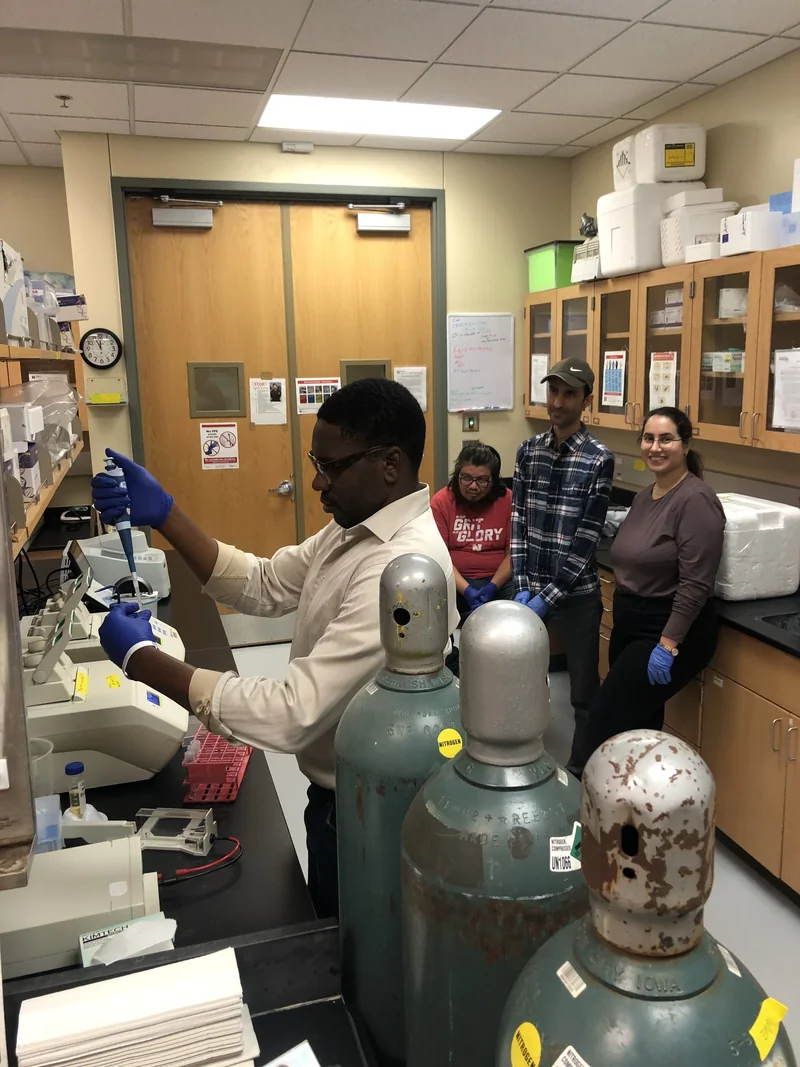 A researcher pipettes at a laboratory bench with three colleagues behind him and gas cylinders in the foreground.