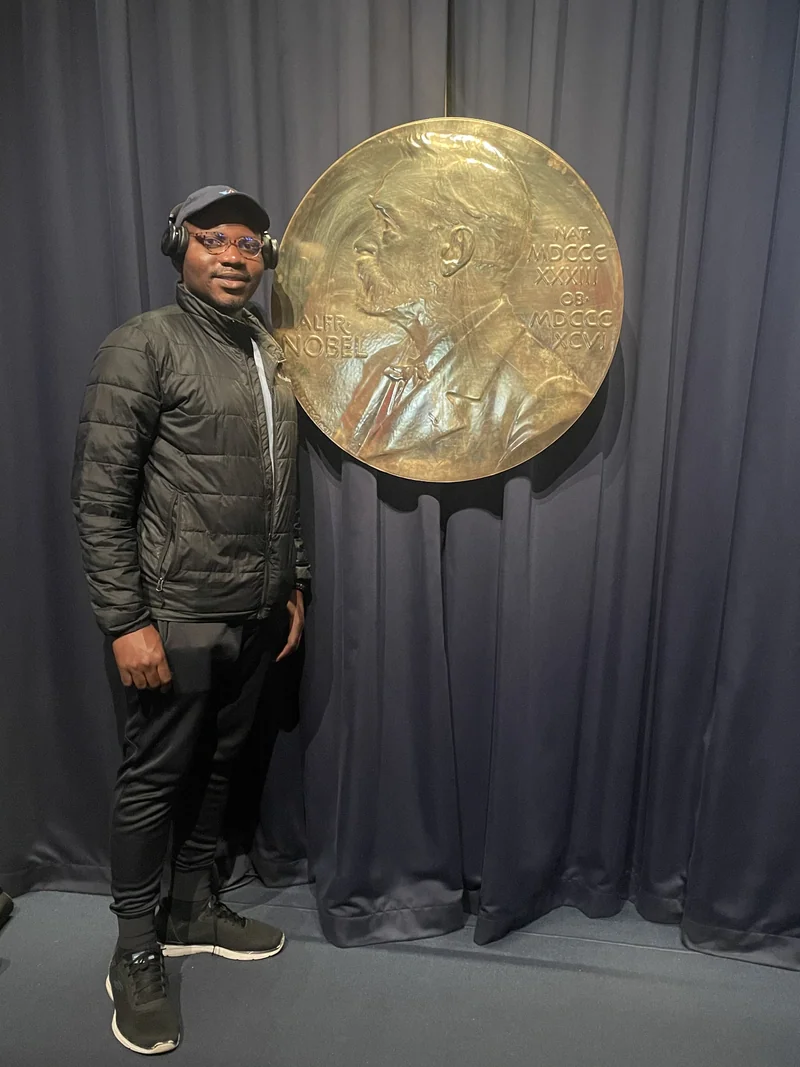 A museum visitor in a dark jacket stands beside a large gold Nobel medal display against a dark curtain.