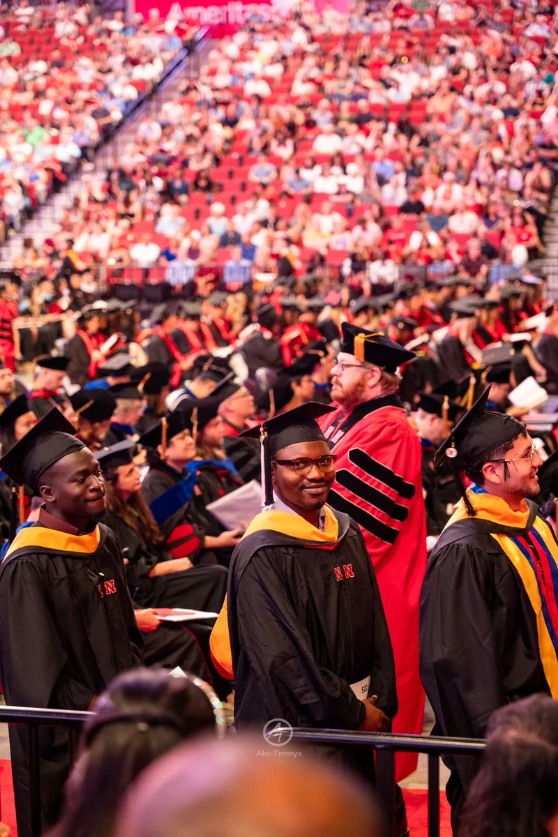 A graduate in black Nebraska regalia and a gold hood stands among graduates and faculty in a crowded arena.