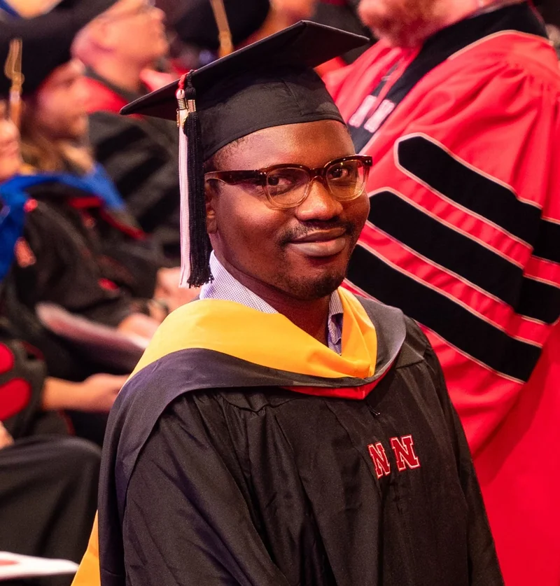 Close-up of a graduate in black Nebraska regalia and a gold hood during an indoor commencement ceremony.